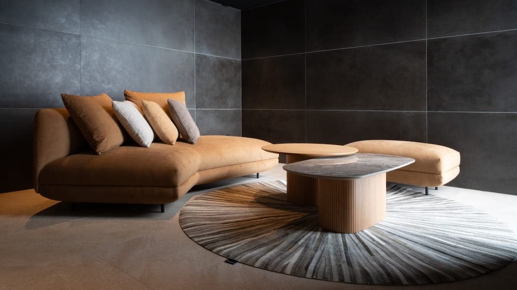 Curved sofa in caramel suede with layered cushions, paired with a round wood coffee table and marble-topped side table on a patterned rug, set against dark stone walls