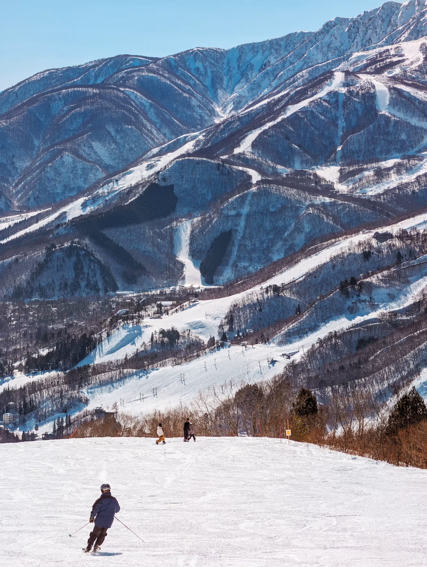 Skier descending a groomed run above Hakuba, with ski trails carved into the mountainside and the valley village below
