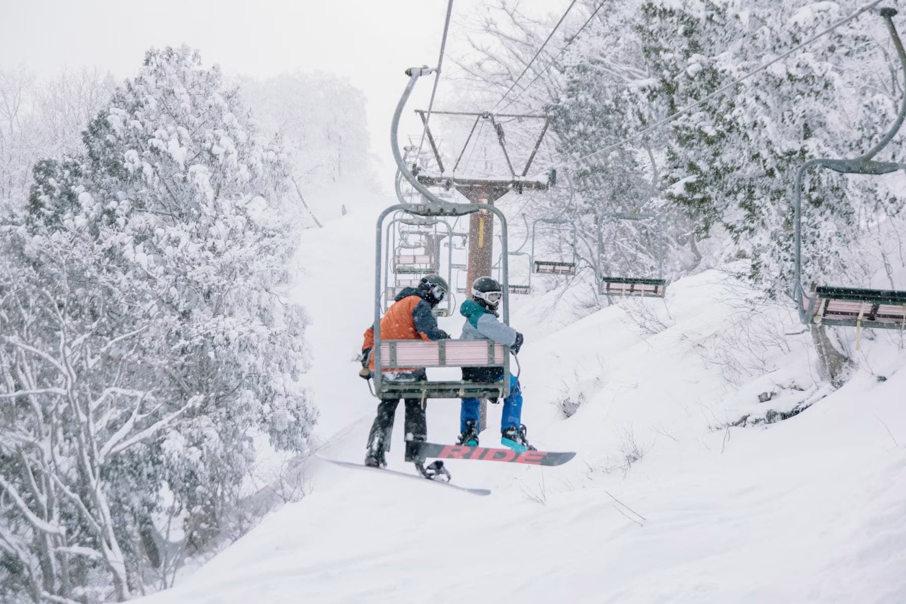 Two snowboarders riding a chairlift through falling snow, surrounded by snow-laden trees in Hakuba
